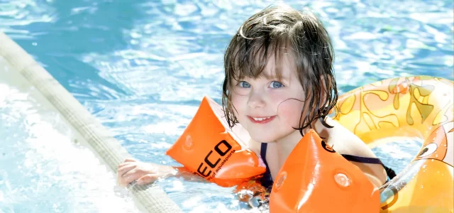 Mädchen mit Schwimmflügeln und Schwimmreifen am Beckenrand im Wasser