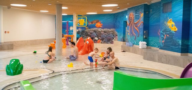 Mehrere Familien sitzen und spielen gemeinsam im seichten Wasser eines Kinderbeckens mit bunten Unterwasser-Wandmalereien und Spielelementen.