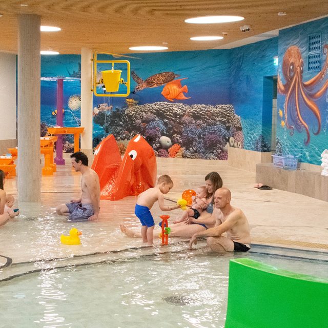 Mehrere Familien sitzen und spielen gemeinsam im seichten Wasser eines Kinderbeckens mit bunten Unterwasser-Wandmalereien und Spielelementen.