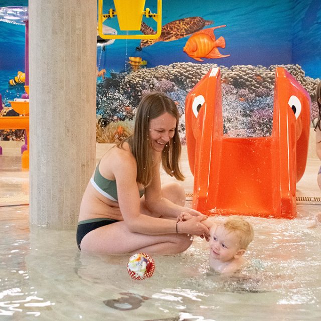 Eine Frau sitzt im flachen Wasser und spielt mit einem Kleinkind, während ein weiteres Mädchen neben einer kleinen orangefarbenen Elefantenrutsche steht.
