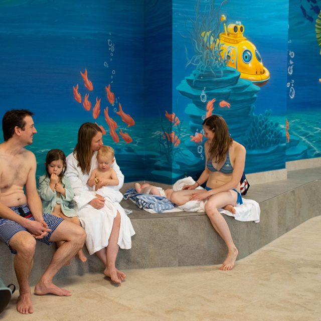 Drei Erwachsene und drei Kleinkinder sitzen gemeinsam auf einer Bank vor einer Unterwasser-Wandmalerei und entspannen sich.