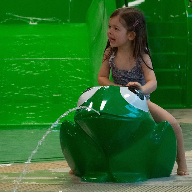 Ein kleines Mädchen sitzt lächelnd auf einer grünen Froschfigur, die Wasser spritzt, vor einer großen grünen Rutsche.