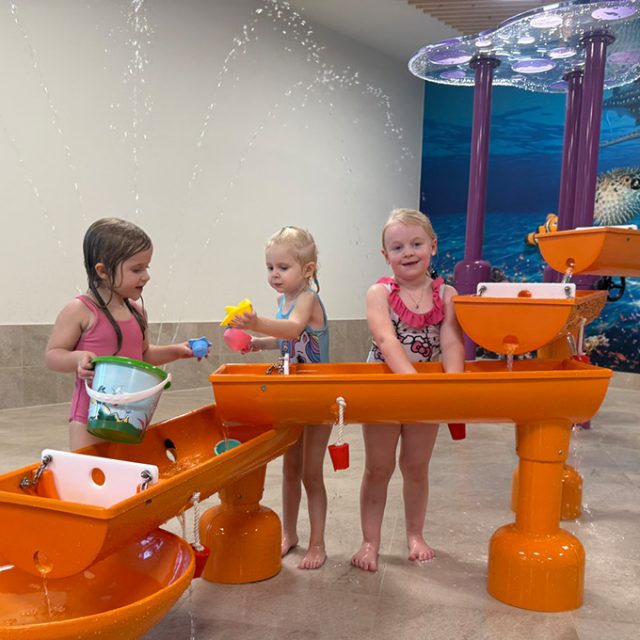 Drei kleine Mädchen spielen fröhlich an einer orangefarbenen Wasserspielanlage mit Eimern und Rinnen.