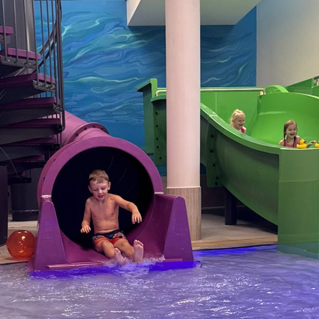 Ein Junge rutscht aus einer lila Röhrenrutsche ins Wasser, während zwei Mädchen in der grünen Breitrutsche im Hintergrund warten.