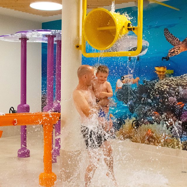 Ein Mann hält ein Kleinkind in einem Wasserspielbereich, während sie von einem kippenden Wassereimer nass gespritzt werden.