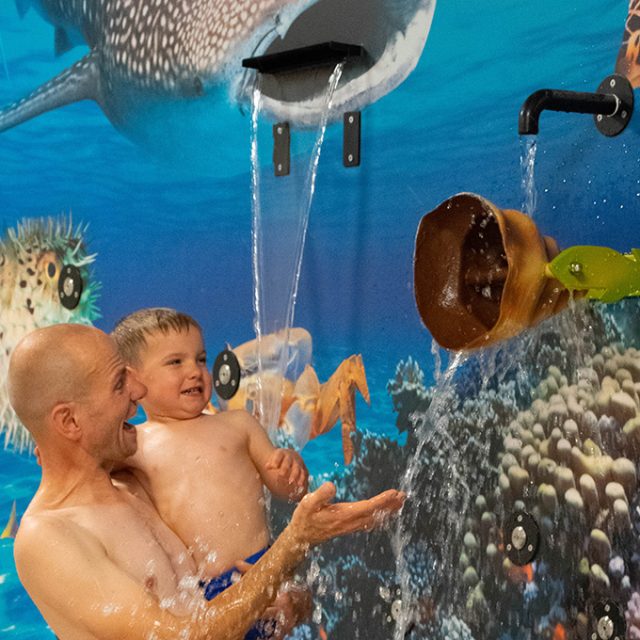 Ein Mann und ein Kleinkind lachen gemeinsam, während sie vor einer Wand mit Unterwasser-Wandmalerei und verschiedenen Wasserspielelementen stehen.
