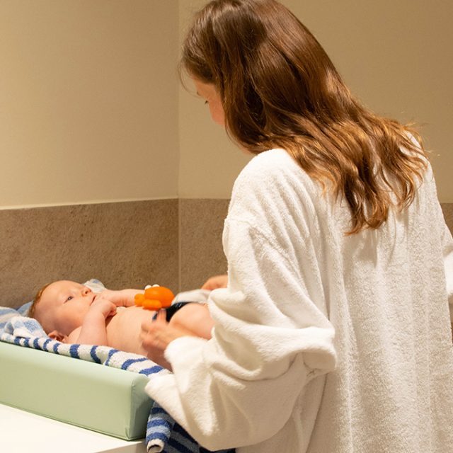 Eine Frau in einem Bademantel wickelt ein Baby auf einer Wickelunterlage in einem hellen Wickelbereich.