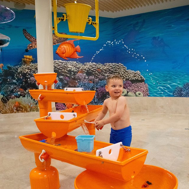 Ein fröhlicher Junge plantscht an einem orangefarbenem Goldwaschrinne mit einer Gießkanne und Eimer.