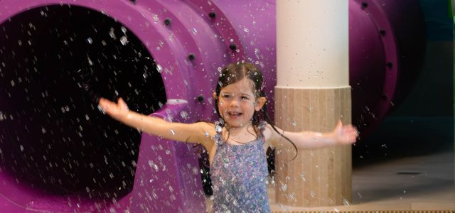 Ein kleines Mädchen im Badeanzug steht lachend und mit ausgebreiteten Armen vor einer lilafarbenen Wasserrutsche, während Wasser spritzend um sie herum fällt.