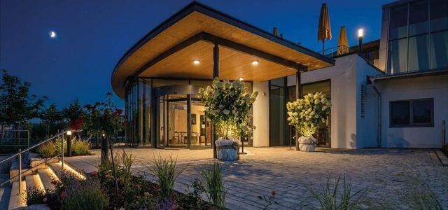 Der Eingangsbereich der Therme Obenrsees bei Nacht, umgeben von bepflanzten Beeten unter einem klaren Sternenhimmel mit einer Mondsichel.